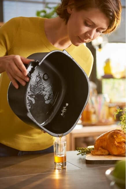 Woman pouring oil from Philips Airfryer XXL pot into a glass on a kitchen table with cooked chicken.